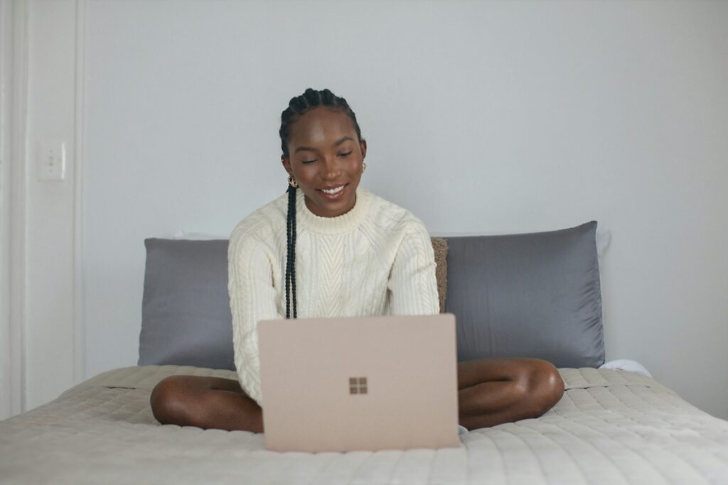 A young Black student researches on her laptop as she sits on her bed.
